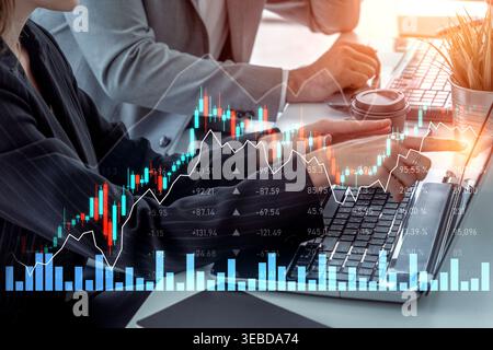 Two business professionals analyze financial data with digital graphs and charts on laptops in a modern office, highlighting teamwork and decision Stock Photo