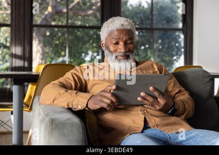 Senior man sitting on gray sofa at home using tablet in protective folio case, wearing smartwatch Stock Photo
