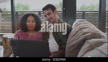 Watching couple on beige sectional sofa sharing laptop in lounge, wearing rose top and olive jacket Stock Photo