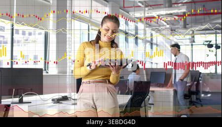 Using tablet with stylus, woman in mustard top and beige pants analyzing charts in open-plan office Stock Photo