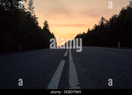 Empty highway road leading through a dense forest under a vibrant orange sunset sky Stock Photo