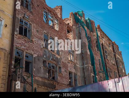 Damaged urban brick building with shattered windows and crumbling walls featuring green safety netting under blue sky Stock Photo