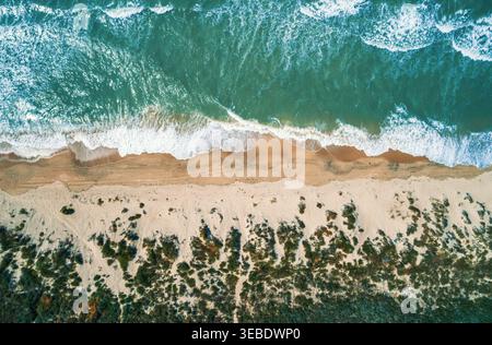Aerial view shows vibrant green ocean water with white foam waves crashing onto a tan sandy beach. Stock Photo
