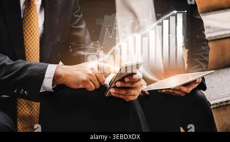Two business professionals analyze financial growth data on a smartphone and tablet, surrounded by digital graphs and charts, highlighting their Stock Photo