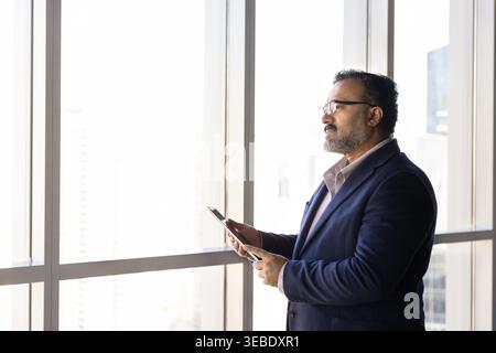 Thoughtful businessman, company executive standing by office window with tablet Stock Photo
