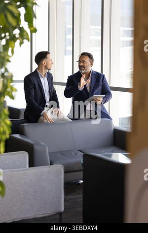 Two businessmen sitting in office lounge, discussing information during workday Stock Photo