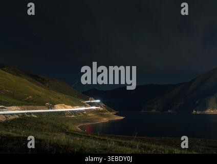 Vehicle light trails glow along a winding mountain road next to a serene lake under a dark night sky Stock Photo