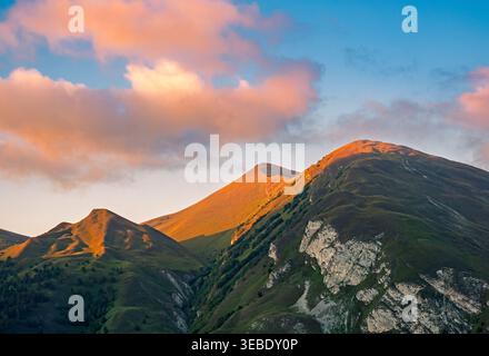 Mountain peaks bathed in warm golden light under a sky with soft orange clouds at an early morning or late evening Stock Photo