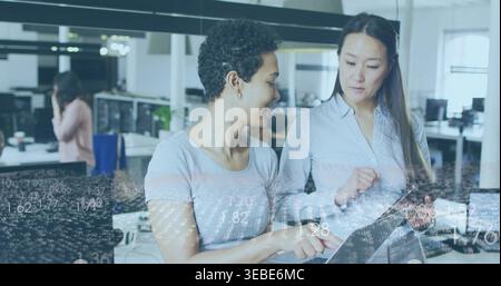 Collaborating women wearing grey tee and pale blouse reviewing tablet at open office, data overlay Stock Photo