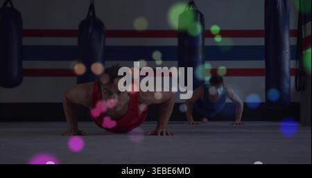 Performing pushups front man wearing red tank top on gym mat, with punching bags Stock Photo