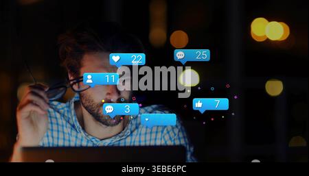 Holding glasses, man in blue checked shirt working on laptop at desk at night, floating alerts Stock Photo