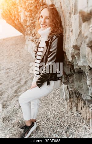 Woman beach fashion smiling woman in black leather jacket and white pants leaning on a rock at pebble beach Stock Photo