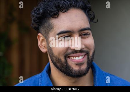 Man in his 30s smiling and looking right in yard wearing blue denim jacket with beard Stock Photo