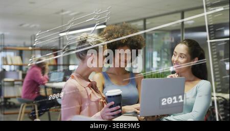 Collaborating three adult women typing on laptop in modern study area, holding travel coffee cup Stock Photo