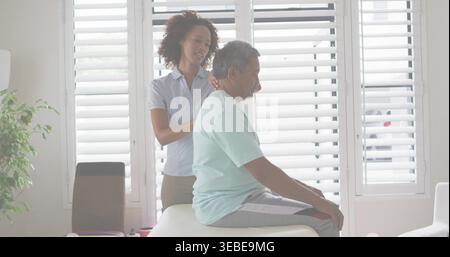 Massaging caregiver wearing blue blouse standing behind senior man on exam table at home, shutters Stock Photo