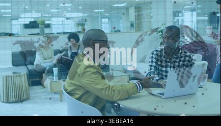 Talking two men, mustard-jacket, using laptop on round-table at office lounge with dotted map panel Stock Photo