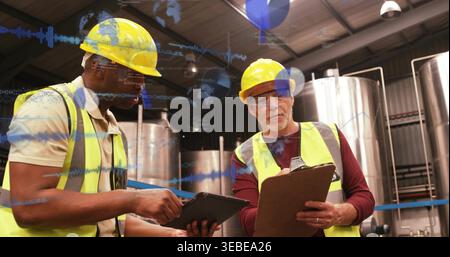 Inspecting two men in yellow hard-hats, safety-vests checking tablet in processing plant Stock Photo