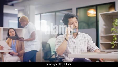 Talking midadult Asian man wearing light plaid shirt with rolled sleeves at open office, on phone Stock Photo
