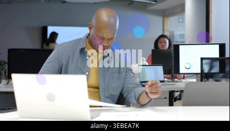Reviewing bald man in shirt typing on laptop, scanning documents at open office with monitors Stock Photo