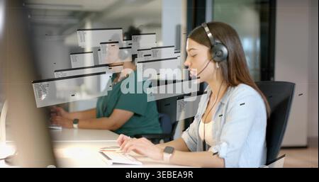 Speaking Indian woman at right typing on keys in office with light-blue shirt and headset, monitor Stock Photo