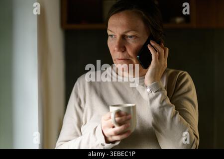 Thoughtful middle-aged woman sips coffee while speaking on her smartphone in a dim kitchen, conveying quiet concentration, morning routine and private Stock Photo
