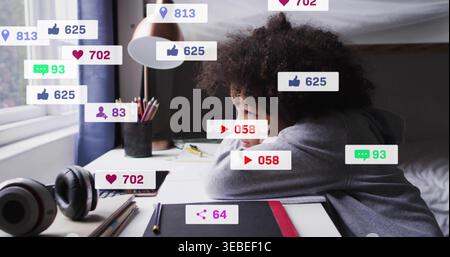 Leaning boy in grey hoodie resting on arms at bedroom desk, with tablet and notification overlays Stock Photo