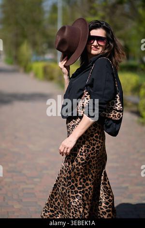 Woman walking in park holding hat and wearing sunglasses, stylish outfit with leopard skirt and black top, woman moving along path in sunlight, outdoo Stock Photo