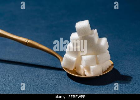 Pile of sugar cubes in spoon on blue background. High stack of white refined sugar cubes in golden spoon on blue textured surface. Concept of sugar ad Stock Photo