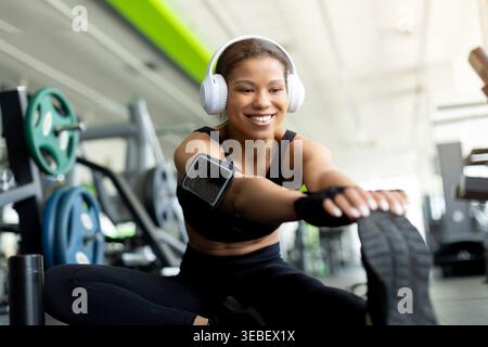 Smiling young Black woman doing seated leg stretch on gym floor Stock Photo