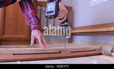A worker in casual clothes uses a screwdriver to tighten fasteners on the side panel of a light wood bed on a dismantled wooden floor in a room Stock Photo