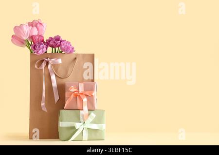 Paper bag with beautiful tulips and gift boxes on yellow background. Mother's Day celebration Stock Photo