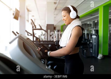 Happy Black woman adjusting treadmill in gym, holding water bottle and smiling Stock Photo
