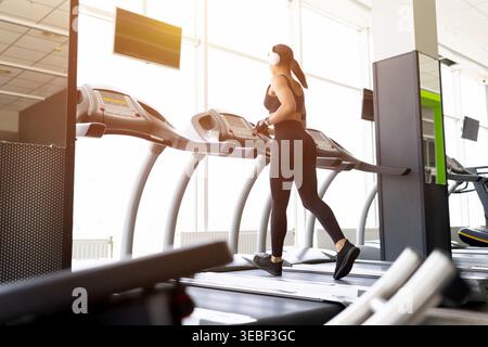 Young Black woman running on treadmill in gym, wearing white headphone Stock Photo