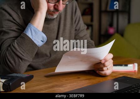 Mature man sitting at table reviewing bills with worried expression. Home interior with laptop and documents. Warm light, horizontal composition, fina Stock Photo