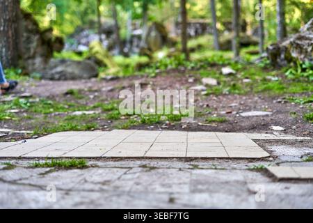A low-angle close-up view of a paved stone pathway transitioning into a dirt forest trail. Light-colored square paving tiles arranged in a grid Stock Photo