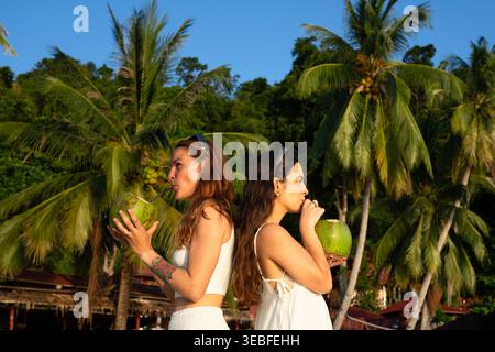 Two women enjoying fresh coconuts on a sunny tropical beach with palm trees Stock Photo