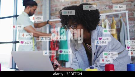 Leaning female designer in gray blazer and glasses, using laptop and smartphone near clothing rack Stock Photo