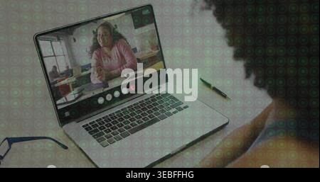 Watching presenter in pink sweater smiling on laptop screen with meeting UI at desk, eyeglasses pen Stock Photo