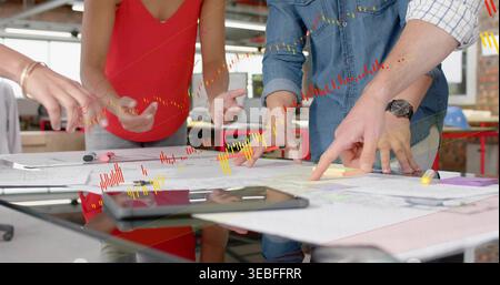 Reviewing three designers in work shirts leaning over plans in studio with tablet and blue helmet Stock Photo