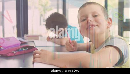 Smiling boy in grey shirt yellow-trim leaning on table holding pink pencil purple case in classroom Stock Photo