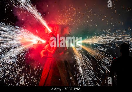 Night festival scene with performers carrying fireworks emitting bright sparks and smoke, dramatic long exposure light trails and silhouetted onlooker Stock Photo