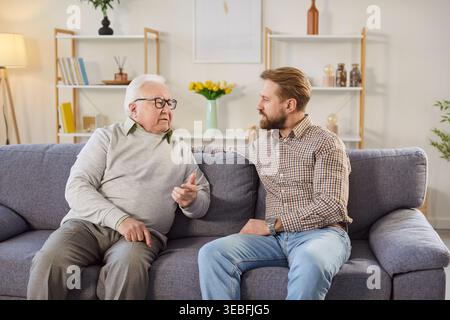 Serious senior father talking with adult son while sitting on sofa and sharing thoughts at home. Stock Photo