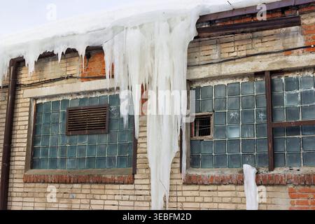 Massive icicles hang from the old brick building's roof overhang, framing teal glass block windows and signs of decay Stock Photo