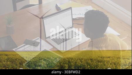 Working woman in yellow sweater typing on laptop at home with wind turbines overlay, copy space Stock Photo