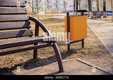 Yellow metal trash bin in a city park next to a brown wooden bench and a pedestrian path. Municipal metal trash container for combat litter and mainta Stock Photo
