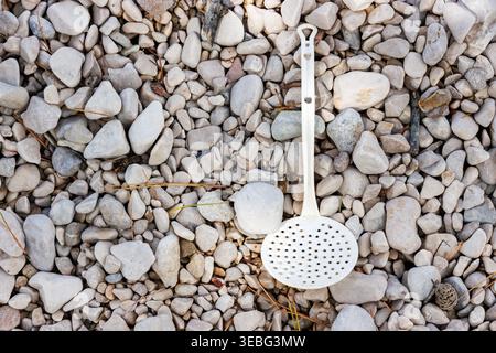 Discarded slotted spoon on pebble beach background top view. Plastic waste and beach pollution with copy space. Stock Photo