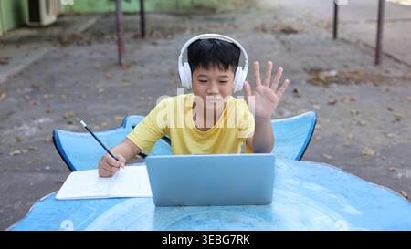Cute Asian boy wearing headphones, waving hand to a laptop camera while studying online. Concept of virtual classroom, video conference, and digital c Stock Photo