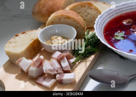 Beetroot soup with lard and bread on a wooden board. Traditional Slavic meal served in a white bowl. Healthy homemade lunch with vegetable broth and rye bread. Culinary concept for menu design. Stock Photo