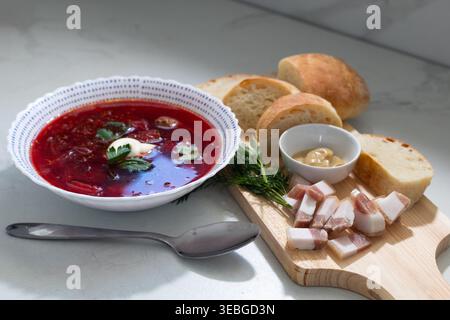 Bowl of traditional borscht soup topped with sour cream and fresh dill garnish. Served with rustic bread slices and smoked bacon on wooden Stock Photo