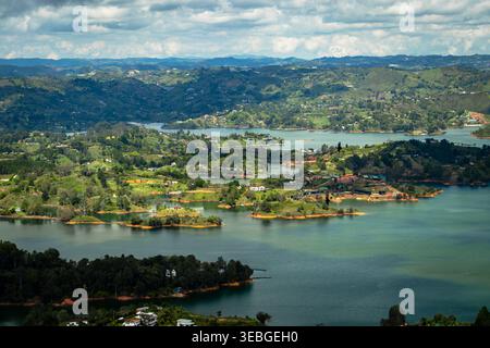 Wide aerial view of the Peñol-Guatapé reservoir with tranquil water, lush green hills, small islands, and scattered homes under a cloudy sky. Peaceful Stock Photo